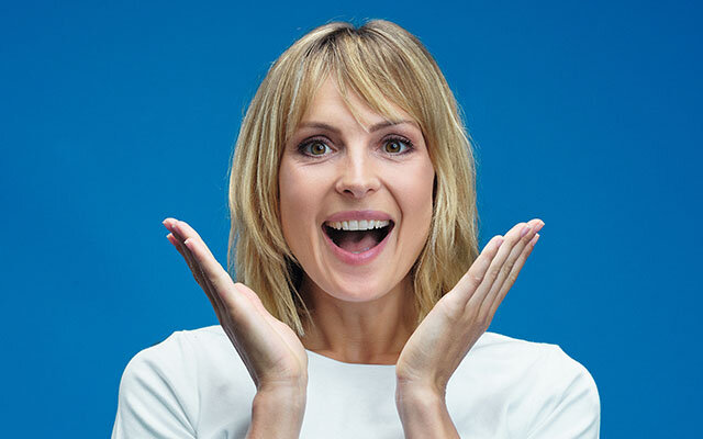 Eine blonde Frau mit braunen Augen steht vor blauem Hintergrund und hält sich aufgeregt die Hände neben das grinsende Gesicht.