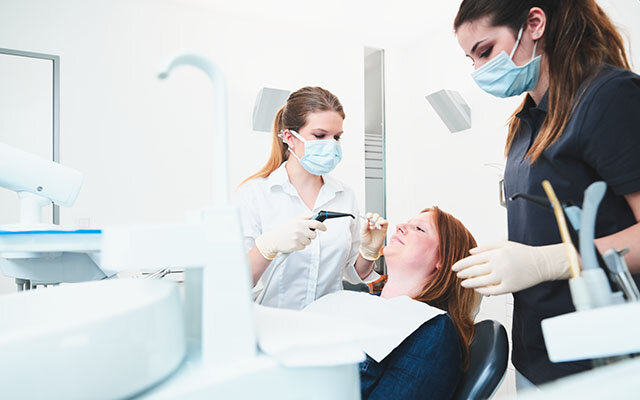 Eine rothaarige Frau liegt auf einem Zahnarztstuhl und wird von einer blonden Zahnärztin und einer dunkelhaarigen Zahnarzthelferin behandelt.