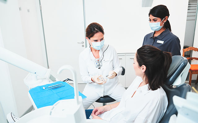 Patientin sitzt im Behandlungszimmer auf dem Behandlungsstuhl und bekommt etwas von der Zahnärztin erklärt, ihre Assistentin steht daneben