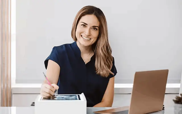 Lächelnde Frau sitzt am Schreibtisch mit Laptop und macht Notizen.