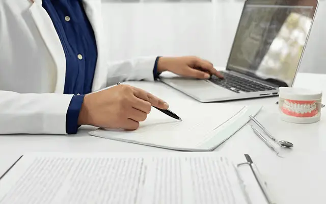 Zahnärztin macht Notizen am Laptop neben zahnmedizinischen Instrumenten.