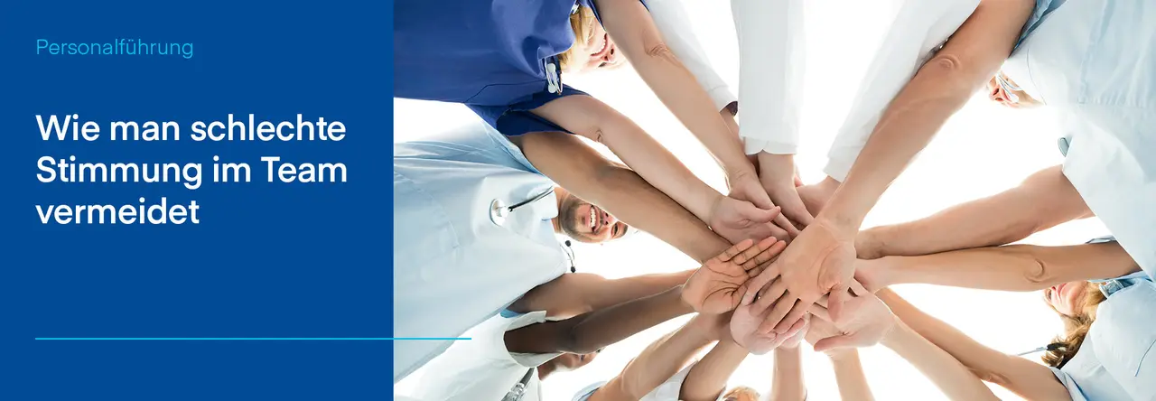 Mehrere Hände, die übereinander gestapelt sind, bilden einen Kreis und symbolisieren Teamgeist und Zusammenhalt. Die Menschen tragen teilweise medizinische Kleidung, was auf ein Gesundheitsteam hindeutet. Links im Bild steht auf einem blauen Hintergrund der Text: 'Wie man schlechte Stimmung im Team vermeidet'
