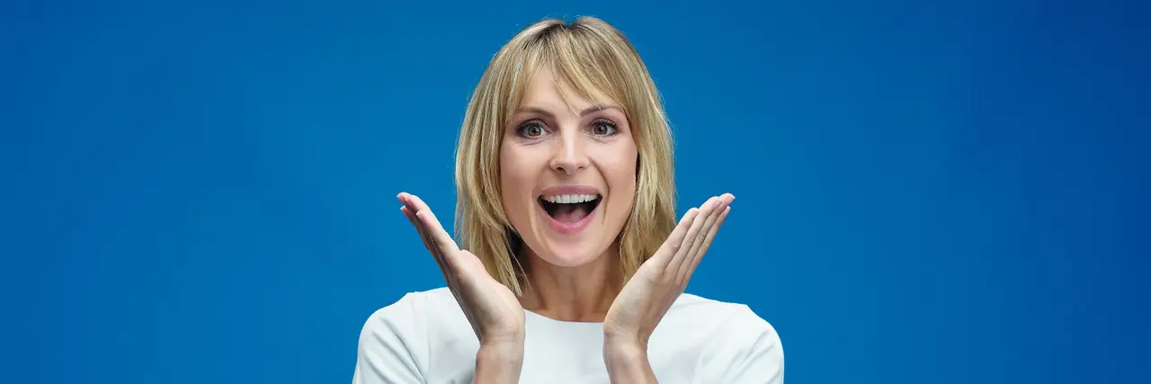 Eine blonde Frau mit braunen Augen steht vor einem blauen Hintergrund und hält sich die Hände grinsend neben den Mund.